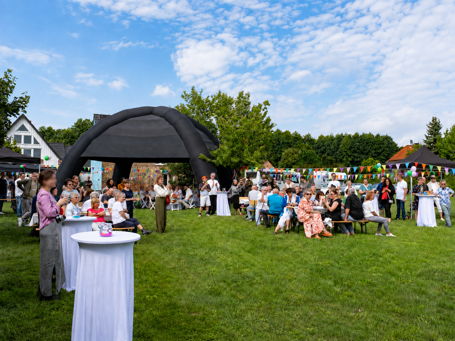 Gartenfest auf der Wiese: Gäste an Stehtischen und Bierbänken unter Girlanden vor dem Pavillonzelt